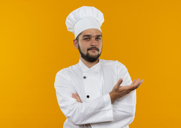 厨师自信的年轻男性厨师身着厨师制服 站在封闭的姿势下 在橘色的墙上展示着孤立的空手 还有复制空间年轻男性制服
