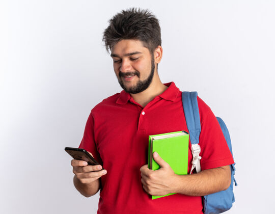 个人快乐的年轻留着胡子的学生 穿着红色马球衫 背着背包 拿着笔记本 拿着智能手机 站在白色的墙上兴高采烈地笑着看着它年轻人手机欢呼