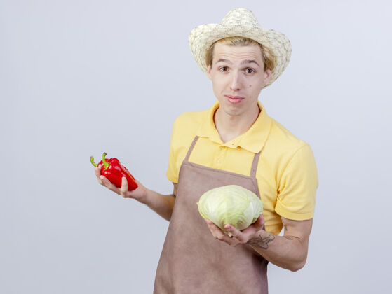 胡椒年轻的园丁穿着连体衣 戴着帽子 手里拿着卷心菜和红甜椒微笑着年轻花园抱着
