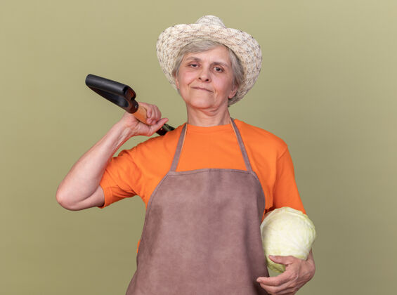 园艺高兴的上了年纪的女园丁戴着园艺帽 肩上扛着白菜和铁锹请绿色戴