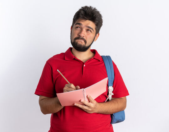 姿势年轻的留着胡子的学生 穿着红色马球衫 背着书包 站在白色的墙上 困惑地抬头 在笔记本上写字笔记本马球胡须