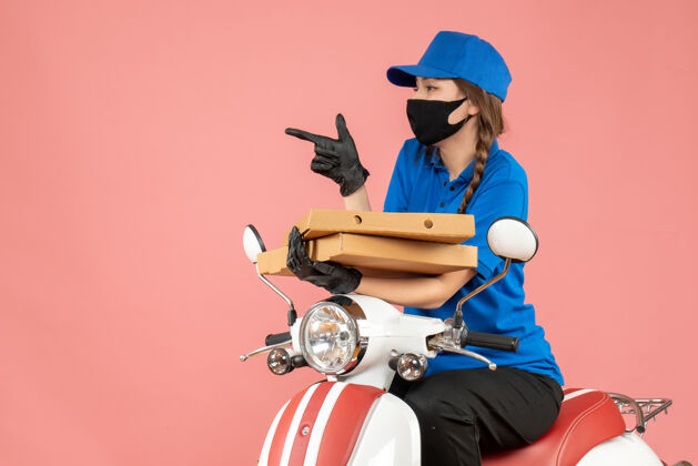 摩托车前视图年轻好奇的女快递员戴着医用面罩和手套坐在滑板车上传递粉彩桃背景的命令秩序滑板车坐着