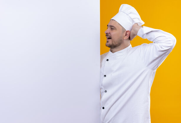 墙穿着厨师制服的年轻帅哥厨师站在白色墙壁后面 用手指着隔离在橙色墙壁上的复制空间印象制服手