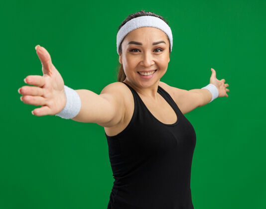 年轻戴着头巾和臂章的年轻健身女士微笑着自信地站在绿色的墙壁上做运动站立自信女人