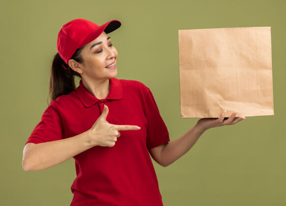 指向年轻的送货员身穿红色制服 戴着帽子 手里拿着纸包 食指指着纸包 自信地微笑着站在绿色的墙上举行立场包装