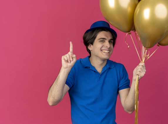 持有面带微笑的白人帅哥戴着蓝色派对帽手持氦气球指向上派对气球氦