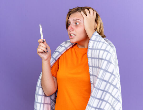 体温计不健康的年轻女子裹着温暖的毯子 感觉不舒服 患流感 发烧 用温度计测量体温 站在紫色的墙上 看起来很担心担心暖和站立