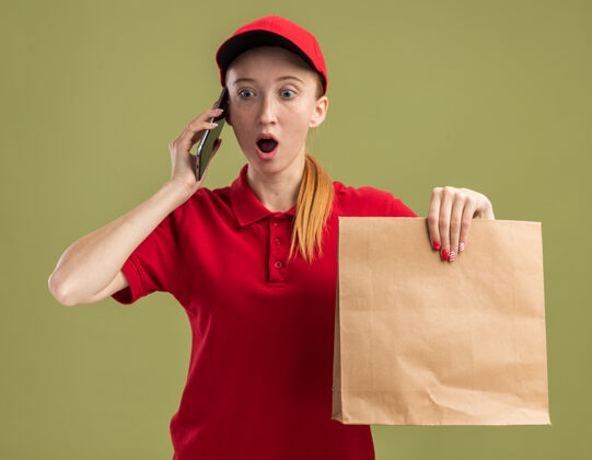 电话年轻的送货员身穿红色制服 戴着帽子 手里拿着一个纸包裹 一边看着绿色的墙壁一边用手机交谈 一边惊讶不已帽子年轻送货