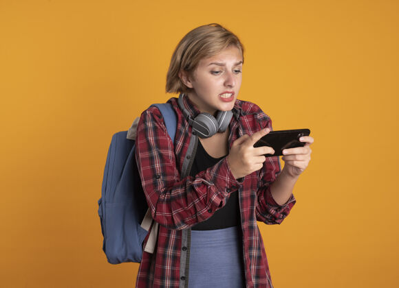 穿着焦急的年轻斯拉夫女学生戴着耳机背着背包 看着手机电话背包橙色