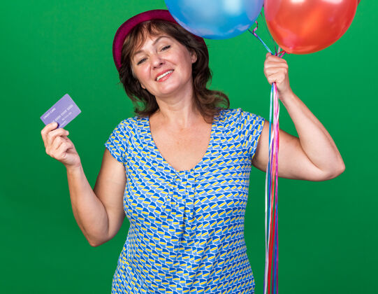 年龄快乐积极的中年妇女 戴着派对帽 手持五颜六色的气球和信用卡 微笑着站在绿色的墙上欢庆生日派对气球立场帽子