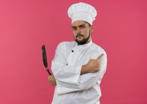 制服自信的年轻男性厨师 身着厨师制服 手拿煎锅 指着粉色墙壁上的隔离面 留有复印空间烹饪信心平底锅