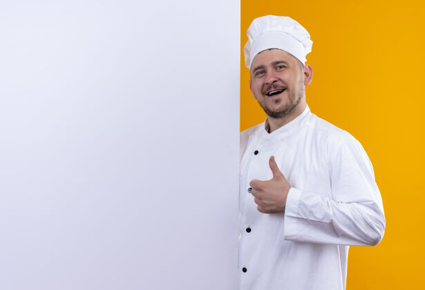 快乐身着厨师制服的年轻帅气的厨师站在白色的墙后 在橙色的墙上孤立地竖起大拇指 留有复印空间墙壁烹饪大拇指