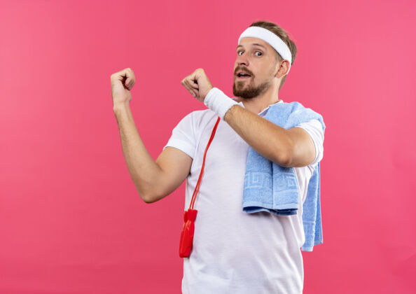 头带令人印象深刻的年轻英俊的运动男子戴着头带和手环指向后面的跳绳和毛巾在肩膀上隔离在粉红色的墙上帅气绳子毛巾