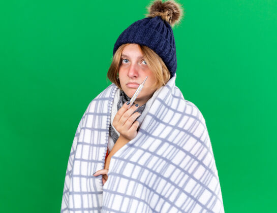 不健康不健康的年轻女子裹着毯子戴着帽子用体温计测量体温患流感发烧站在绿色的墙上女人毯子包裹