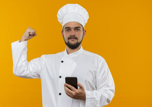 手势自信的年轻男厨师身着厨师制服 手持手机 在橙色的墙壁上做着强烈的手势 并留有复印空间厨师自信手机