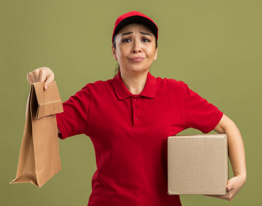 迷茫身穿红色制服 头戴鸭舌帽的年轻女送货员站在绿色的墙上 手里拿着纸包装和纸板箱 感到困惑和不快抱着包装站着
