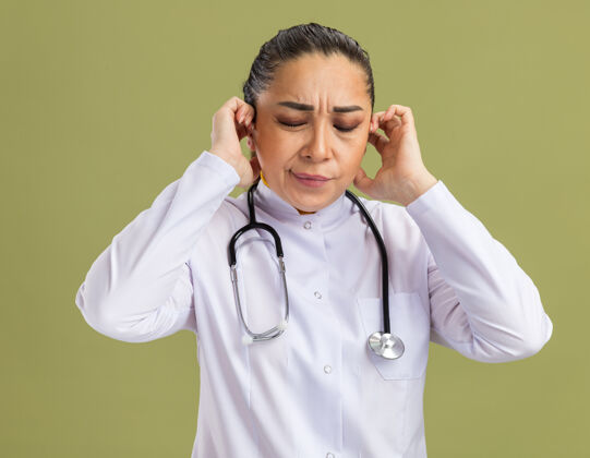 女人年轻的女医生 穿着白药衣 脖子上戴着听诊器 看着不舒服 站在绿墙上摸着头痛的头痛苦触摸年轻
