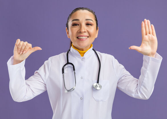 显示年轻的女医生身穿白色药衣 脖子上戴着听诊器 竖起大拇指 张开双臂 自信地站在紫色的墙上年轻微笑外套