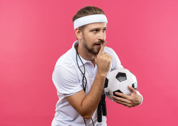 绳子令人印象深刻的年轻英俊的运动男子戴着头带和腕带拿着足球手势沉默和看一边跳绳围绕着他的脖子隔离在粉红色的墙上球手势帅气