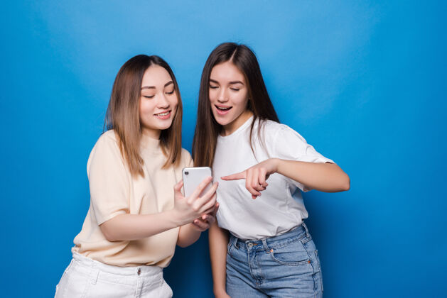 泳衣快乐的两个年轻女孩笑着指着智能手机的屏幕 一边隔着蓝色的墙自拍年轻手指夏天