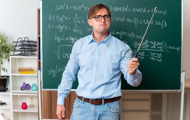 男愤怒的年轻男老师戴着眼镜 拿着指针 站在教室里数学公式的黑板旁讲解功课老师指针戴