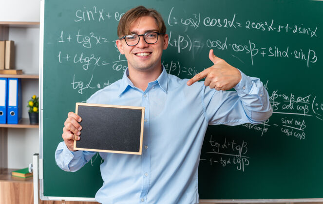 老师年轻的男老师戴着眼镜 拿着小黑板 用食指指着黑板 面带微笑 自信地站在教室里的数学公式黑板旁眼镜近指着