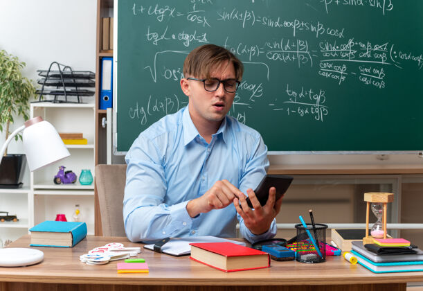 年轻年轻的男老师戴着眼镜用智能手机打字 看起来很自信地坐在课桌旁 教室的黑板前放着书和笔记留言教室人