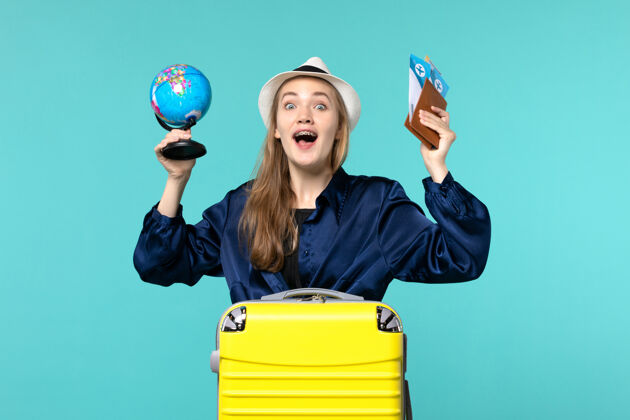小前视图年轻女子拿着机票和小地球仪在淡蓝色背景的飞机上度假远航成人旅程微笑
