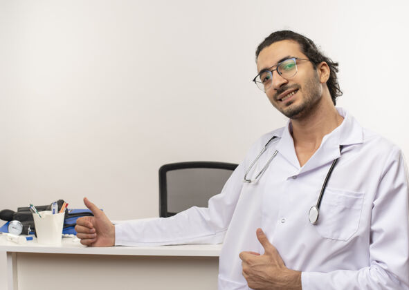 站年轻的男医生戴着医用眼镜 穿着医用长袍 手持听诊器站在办公桌前眼镜副本笔记本电脑
