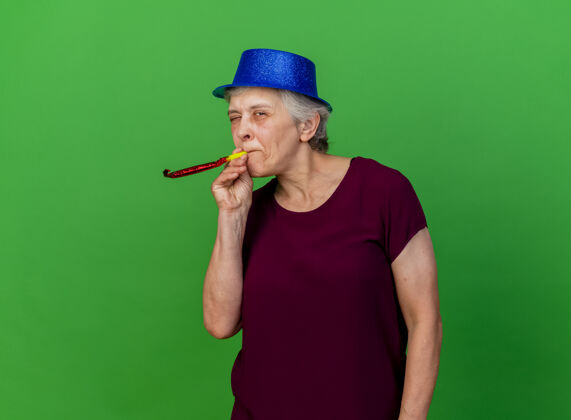 女人戴着派对帽的快乐的老妇人眨着眼睛吹哨子在绿色口哨老人聚会