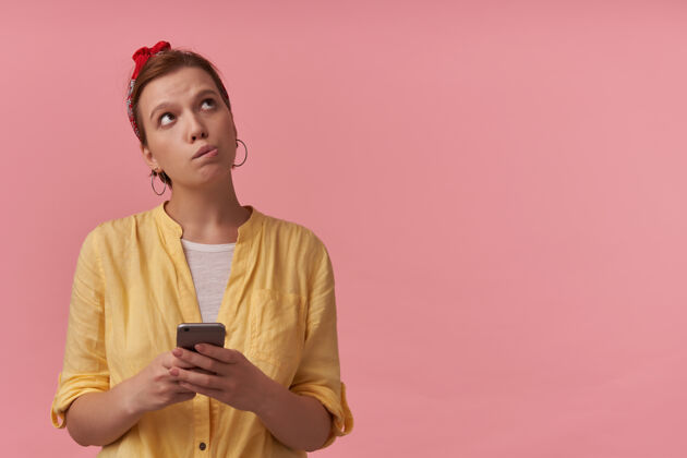 技术沉思迷人的年轻女子 穿着黄色衬衫 头上戴着头带 用智能手机看着粉色墙壁的一边女性衣服壁板