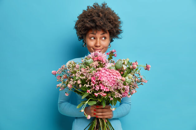 优雅室内拍摄的非裔美国妇女收到美丽的鲜花享受浪漫的约会期待与梦幻沉思的表情旁边得到来自秘密仰慕者或情人的花束隔离在蓝色的工作室墙上人束室内