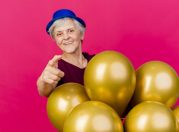 相机微笑着戴着派对帽的老妇人站在氦气球后面 指着粉色的相机粉色气球站着