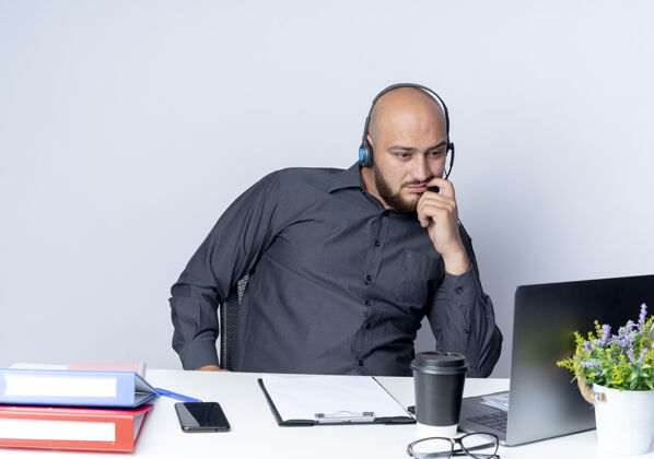 市民体贴的年轻秃头呼叫中心男子戴着耳机坐在办公桌旁 拿着工作工具 看着隔离在白墙上的笔记本电脑笔记本电脑秃头衣服