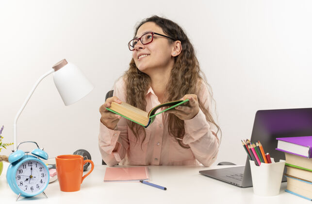 漂亮微笑着的年轻漂亮的女学生戴着眼镜坐在课桌旁 拿着学习工具做作业 拿着书 孤零零地看着白色的墙上作业人感觉
