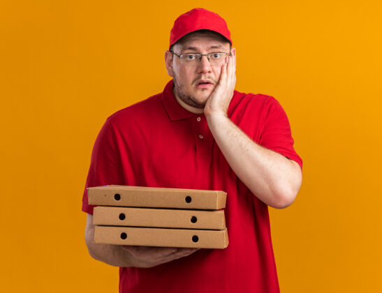 盒子焦急超重的年轻送货员戴着眼镜 手里拿着披萨盒 手放在隔离在橙色墙上的脸上 留着复印空间焦虑孤立比萨饼