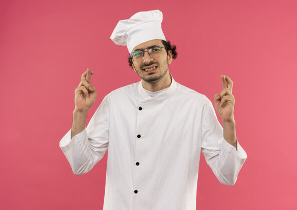 十字架身穿厨师制服 戴着眼镜 手指交叉的年轻男厨师关注背景粉色