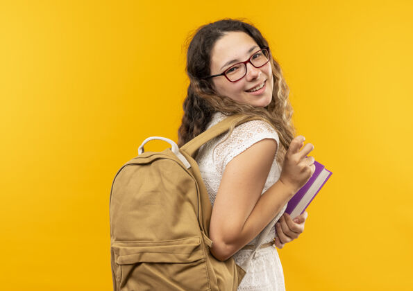 微笑面带微笑的年轻漂亮的女学生站在侧视图中 戴着眼镜 背着书包 隔离在黄色的墙上姿势市民人