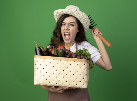 制服年轻的女园丁穿着制服 戴着园艺帽 把菜篮子和耙子孤立地放在绿色的墙上姿势蔬菜表情