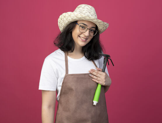 制服微笑着的年轻黑发女园丁戴着眼镜 穿着制服 戴着园艺帽 把锄头耙孤立地放在粉红色的墙上持有人花园