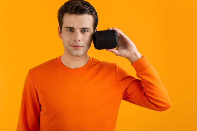 姿势穿着橙色毛衣的时髦微笑的年轻人拿着无线扬声器快乐地听音乐享受橙色的乐趣积极声音乐趣