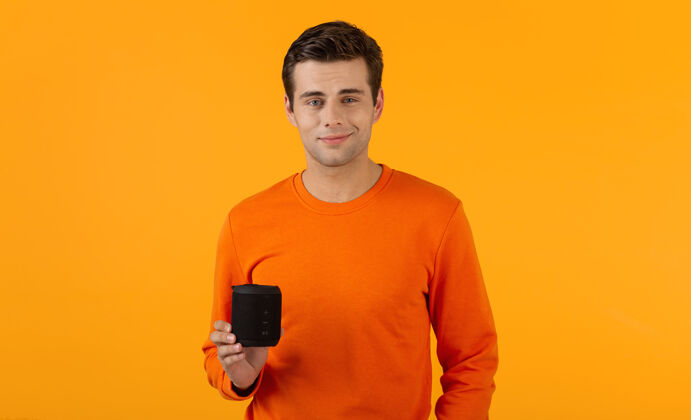 男人穿着橙色毛衣的时髦微笑的年轻人拿着无线扬声器快乐地听音乐享受橙色的乐趣娱乐模特持有