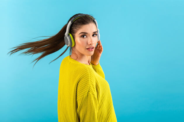 年轻戴着无线耳机听音乐的女人开心地穿着黄色针织毛衣摆着蓝色的姿势可爱时髦姿势