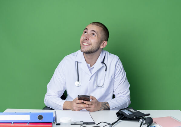 工具面带微笑的年轻男医生 穿着医用长袍 手持听诊器 坐在办公桌旁 手持工作工具 手持手机 孤零零地仰望绿色的墙壁电话桌子人