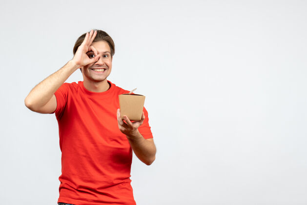 人物一个穿着红色上衣的快乐的年轻人拿着一个小盒子 在白色背景上做着一个戴眼镜的手势红色漂亮肖像
