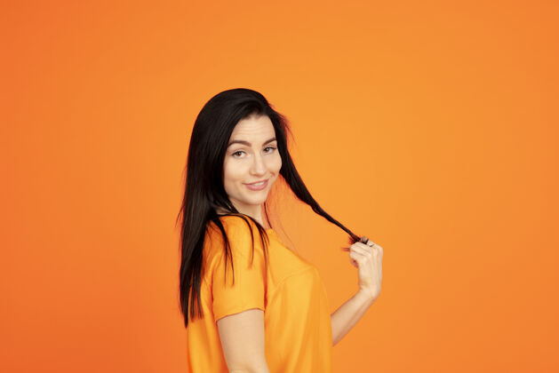 表达橙色工作室背景上的白种人年轻女子肖像穿着衬衫的漂亮深色女性模特人类情感的概念 面部表情 销售 广告广告空间看起来可爱 微笑公司长度面部