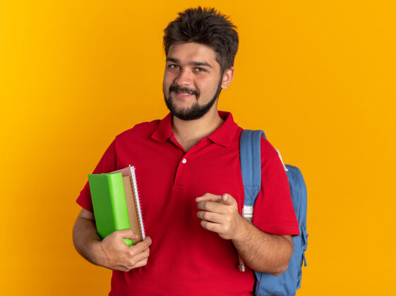 手指年轻的留着胡子的学生 穿着红色马球衫 背着书包 拿着笔记本 食指指着相机 站在橙色背景下 他开心地笑着指着拿着背包