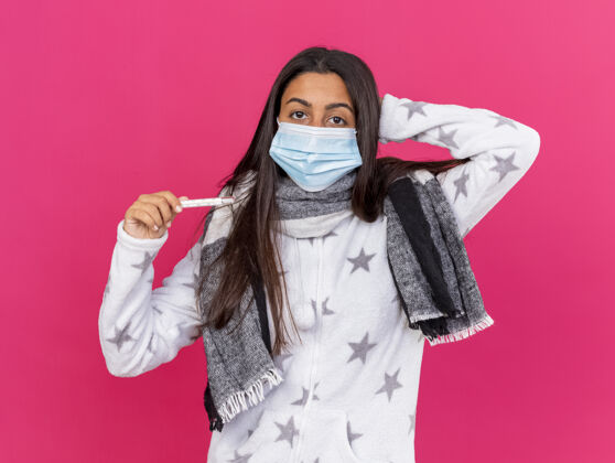 生病困惑的年轻病女孩戴着医用面罩 戴着围巾 拿着体温计 手放在脑后 隔离在粉色上体温计头迷糊
