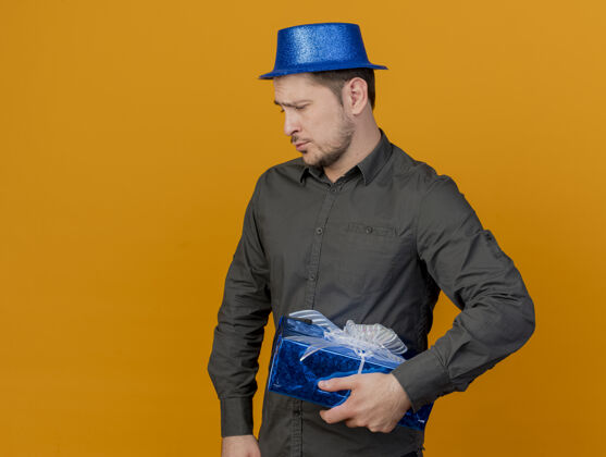 年轻悲伤的年轻人戴着蓝帽子拿着礼品盒孤立在橙色的聚会拿着派对礼物