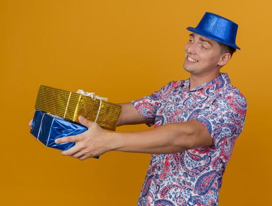 蓝色带着微笑的年轻人戴着蓝色的帽子 拿着礼品盒站在橙色的一边壁板帽子年轻人
